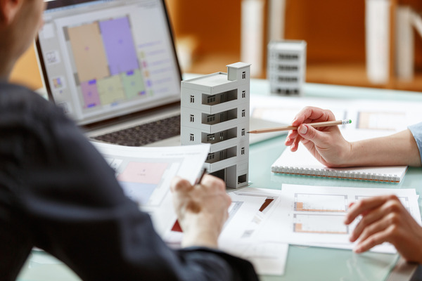 Two designers examine architectural plans and a building model at a table, with a laptop displaying digital layouts in the background.