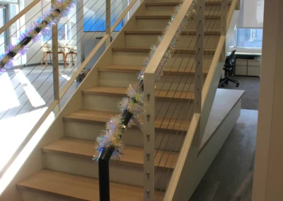 A modern office staircase decorated with colorful tinsel garlands, showcasing wooden steps and sleek metal railings.