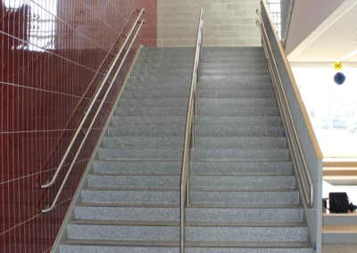 Interior view of a stairwell with gray steps and stainless steel handrails, flanked by red tile and gray block walls. Brightly lit.