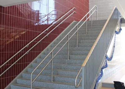 Interior view of a stairwell with gray steps and stainless steel handrails, flanked by red tile and gray block walls. Brightly lit.