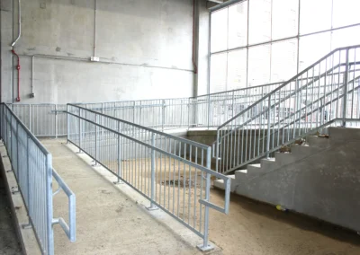 An interior concrete ramp and staircase with metal railings, illuminated by natural light from large windows.