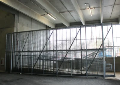 An interior concrete ramp and staircase with metal railings, illuminated by natural light from large windows.