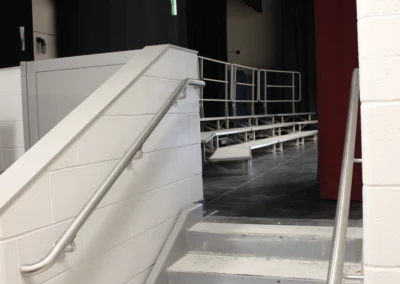 Interior view of a stairwell with gray steps and stainless steel handrails, flanked by red tile and gray block walls. Brightly lit.