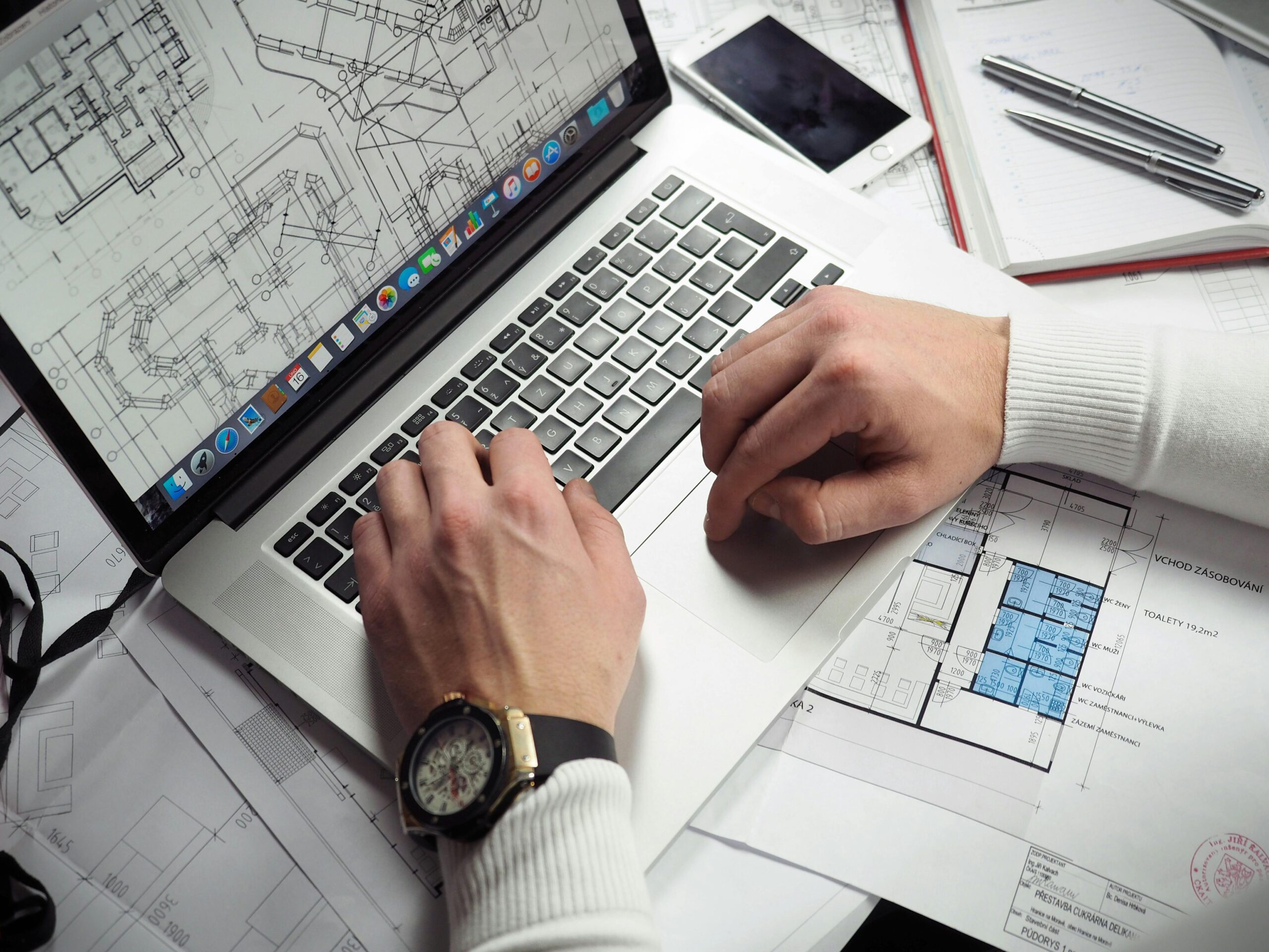 Two designers examine architectural plans and a building model at a table, with a laptop displaying digital layouts in the background.